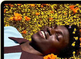 MacBook Neo, color cítrico, persona sonriente tumbada en un campo de flores en la pantalla